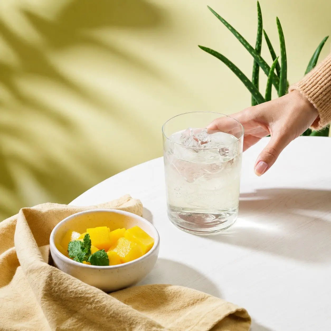 Un verre d'aloe vera glacé posé sur une table claire, à côté d'un bol de mangue et d'un plant d'aloe.